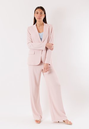 Donna che posa con sicurezza indossando un abito rosa chiaro e una maglietta blu, con capelli lunghi e lisci e un'espressione neutra, su uno sfondo bianco uniforme.