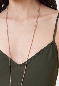 Kaki-grön ärmlös topp med V-ringning, tillverkad av slät tyg, kombinerad med ett fint guldhalsband.