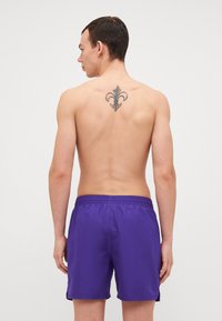 Shorts de bain violets avec une ceinture élastique, texture lisse et fentes sur les côtés ; portés par une silhouette masculine avec un tatouage dans le dos.