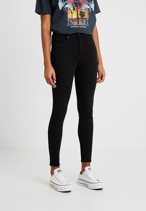 Persoon draagt zwarte skinny jeans, een grafisch T-shirt met vogels en palmbomen, en witte plateau sneakers, staand op een effen witte vloer.