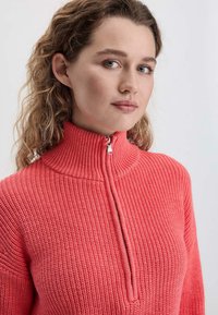 Korallenfarbener Strickpullover mit einem hohen Kragen und halbem Reißverschluss. Verfügt über ein strukturiertes Muster und gerippte Designs an Ärmeln und Kragen.