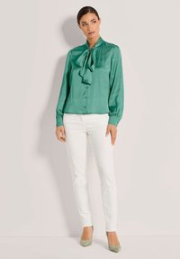 Grüne Satinbluse mit einem gebundenen Kragen, knöpfligem Vorderteil und langen Ärmeln. Kombiniert mit weißen Slim-Fit-Jeans und hellgrünen Absätzen.