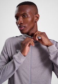 Grå atletisk zip-up jakke med en tekstureret finish, kontrasterende mørke accenter på skuldrene og et logo på brystet.