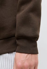 Ermekanten på en brun strikkegenser på en persons arm, kombinert med hvite bukser med tynne, vertikale striper.