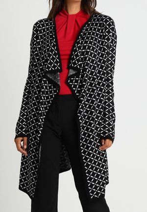 Frau trägt eine lange schwarz-weiß karierte Strickjacke über einer roten, plissierten Bluse und schwarzen Hosen, steht vor einem weißen Hintergrund.