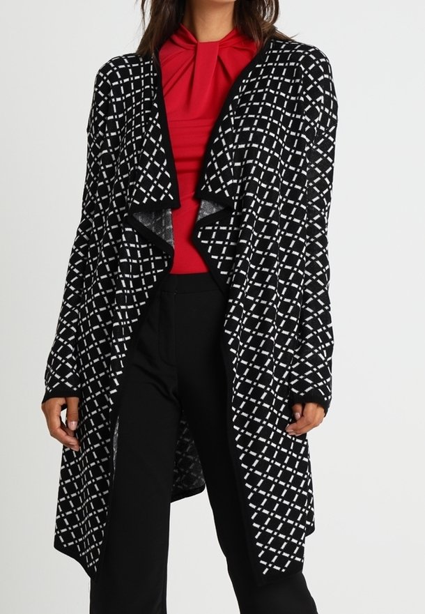 Femme portant un long cardigan à carreaux noir et blanc sur un chemisier plissé rouge et un pantalon noir, debout devant un fond blanc.