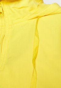 Gul lättviktsjacka med en texturerad huva, synliga sömmar och dragsko-detalj. Slät tyg med en livlig, enhetlig färg.