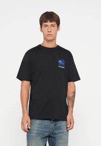 Zwarte katoenen t-shirt met een losse pasvorm, voorzien van een blauwe grafische afbeelding van een kat op de linkerborst en korte mouwen.