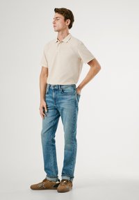 Camisa polo beige, jeans de mezclilla azul, zapatos marrones. La polo tiene un cuello con solapa, los jeans son ligeramente ajustados y los zapatos tienen acentos en contraste.