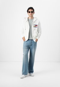 Biely zipsový mikina s nášivkou, šedé tričko, svetlomodré rozšírené džínsy a biele tenisky. Model má tiež na sebe okrúhle slnečné okuliare.