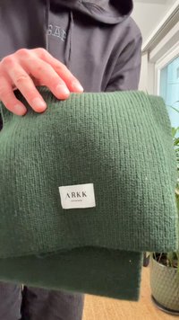 Tamsiai žalia gofruota tekstilė su lygiu paviršiumi, apačioje su balta ARKK etikete. Ranką laiko audinį. Fone matyti augalas.