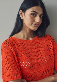 Lys orange hæklebluse med en løstsiddende pasform, der har åbne mønstre og korte ærmer. Udhulningen er bred og ribbet.