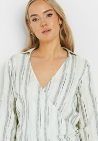 Blusa envolvente ligera con rayas verticales en negro y gris sobre un fondo blanco. Escote en forma de V con tejido texturizado. Mangas largas.