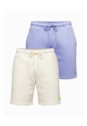 Homme avec barbe portant un sweat crème et un short, des chaussettes blanches avec des rayures rouges et des baskets blanches, debout sur un fond gris uni.