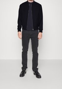 Giacca bomber nera con zip, indossata sopra una camicia scura, abbinata a jeans grigio scuro e stivali neri con lacci. Tessuti di un aspetto liscio.