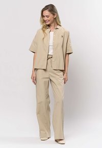 Beige kortärmad skjorta med knappar tillsammans med matchande vida byxor, med en vit linne under och en liten blommig detalj.