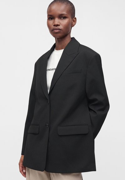Femme portant un blazer noir oversize sur une chemise blanche avec un texte partiel, debout contre un fond clair uni.