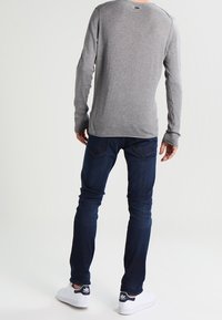 Homme portant une chemise à manches longues grise, un jean bleu foncé et des baskets blanches Adidas, debout de dos sur un fond blanc uni.