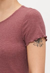 Burgundinė raukšlėta palaidinė su apvaliu iškirpimu, trumpomis rankovėmis ir volangais. Matomas gėlių tatuiruotė ant viršutinės rankos. Minkštas audinio tekstūra.