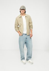 Veste en polaire beige, t-shirt blanc, jeans larges bleu clair et baskets blanches avec des accents gris. Une casquette grise complète le look.