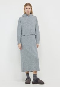 Donna che indossa un felpa a maglia grigia e una gonna lunga abbinata, con calze grigie scure e pantofole, in piedi contro uno sfondo bianco.