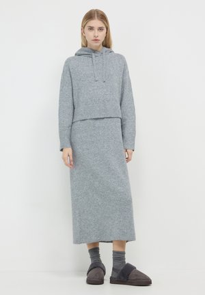 Woman wearing grey knit hoodie and matching long skirt with dark grey socks and slippers, standing against white background.