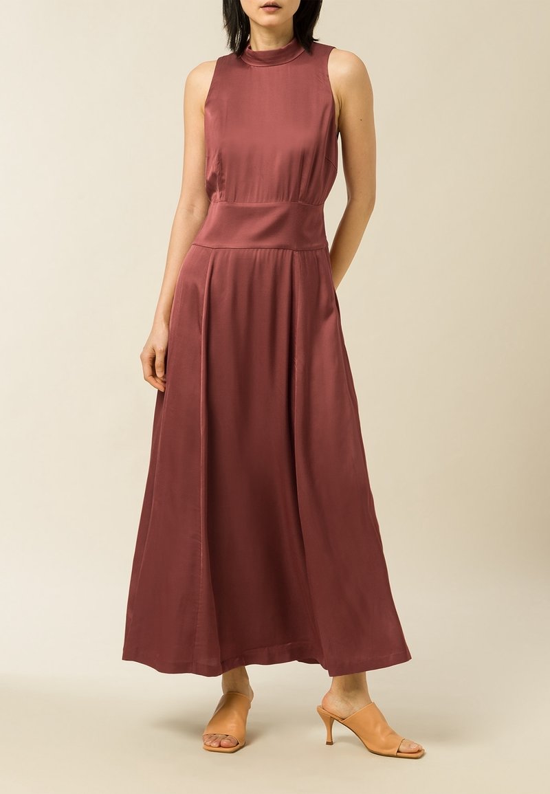 Robe longue sans manches de couleur bordeaux riche, en tissu lisse avec un col haut et une jupe fluide. Portée avec des talons ouverts couleur beige.