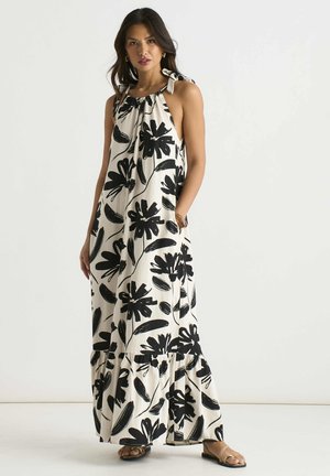 Woman wearing a sleeveless white maxi dress with black floral prints and shoulder tie straps, standing barefoot against a plain background.