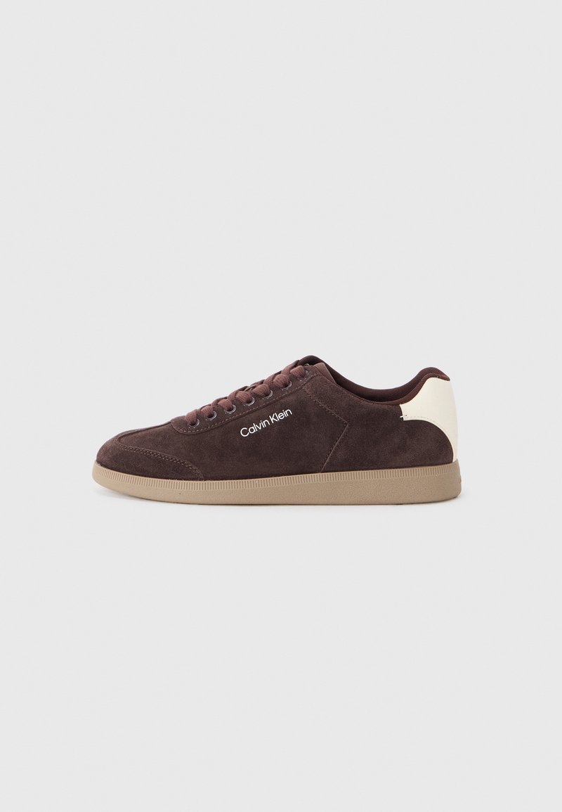 Baskets en suede marron avec un accent de talon beige clair, arborant une marque blanche sur le côté, un bout rond et une semelle en caoutchouc beige.