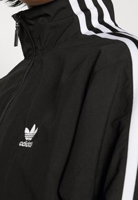 Svart zip-up jacka i lätt material, med vita ränder längs ärmarna och en broderad logotyp på bröstet.