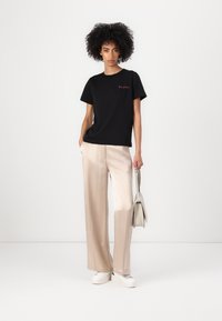 Black cotton t-shirt with "GUESS" logo in red, paired with light beige wide-leg trousers and white sneakers, holding a small beige handbag.
