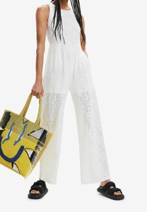 White lace jumpsuit with wide legs and sleeveless design. Paired with a colorful patterned tote bag and black sandals.