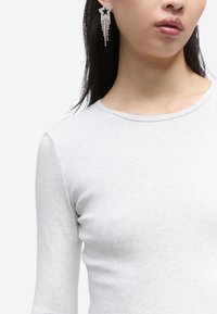 Γυναίκα που φοράει λευκή μπλούζα με μακριά μανίκια και έχει ένα κρεμαστό ασημένιο σκουλαρίκι σε σχήμα αστεριού στο αριστερό αυτί της, φωτογραφημένη από τους ώμους έως το πηγούνι.