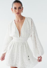 Vit klänning med ett strukturerat, mönstrat ytskikt, djup V-ringning, långa ärmar och ett elastiskt midjeband med dragsko och metallaccenter.