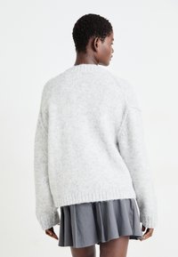 Maglione grigio chiaro oversized con una finitura morbida e testurizzata, orlo a costine e spalle scese, indossato sopra una gonna svasata grigio scuro.