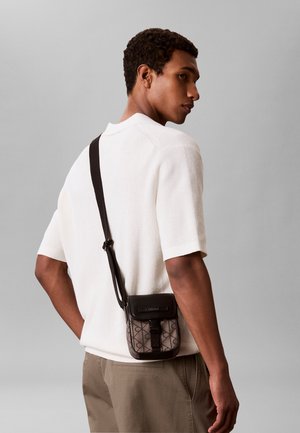 Ung mand iført en hvid kortærmet sweater og beige bukser, bærende en lille mønstret skuldertaske med en sort rem.