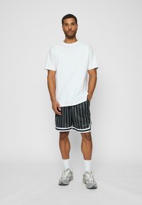 Tricou alb cu mânecă scurtă asociat cu pantaloni scurți negri cu dungi și accente albe, precum și pantofi sport gri cu șosete de culoare deschisă.