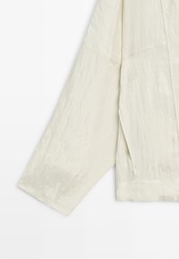 Long sleeve of a cream-colored, slightly textured lightweight jacket with visible seams.