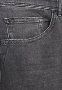 Jean en denim noir avec une poche arrière ornée de détails de couture, un petit clou en métal et une finition de tissu lisse et texturée.