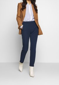 Kvinna i brun kavaj, blårandig skjorta, navybyxor och vita spetsiga ankelskor, stående med ena handen i fickan.