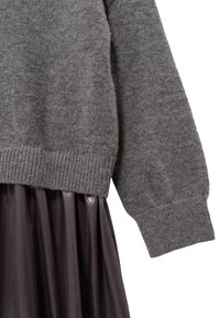 Maglione grigio lavorato a maglia con orlo a coste, abbinato a una gonna plissettata metallizzata scura, che mette in evidenza una morbida texture e dettagli di drappeggio.