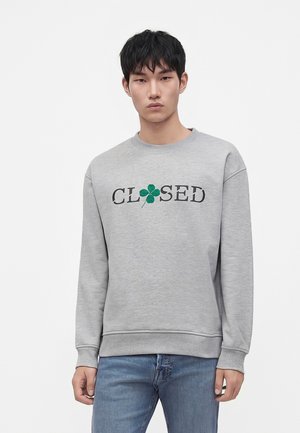 Jeune homme aux cheveux courts et noirs portant un sweat-shirt gris avec le texte "CLOSED" et un trèfle à quatre feuilles vert remplaçant la lettre O, jean bleu.