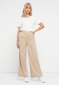 Beige vide bukser med elastisk liv, med forreste folder og sidelommer, parret med en hvid t-shirt med rund hals og hvide sneakers.