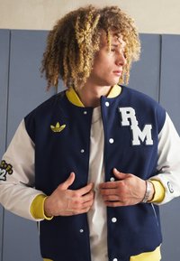 Mornarji in beli šolski jopič z rumenimi poudarki, z vezenjem "RM" in majhnim logotipom Adidas. Mehka tkanina z gumbnim zapiranjem.