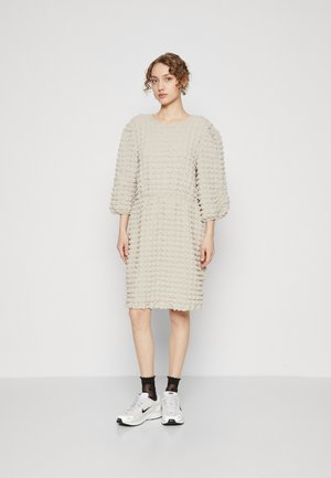 Beige tekstureret kjole med puffede ærmer, knælængde, med en samlet talje og et bølget mønster. Parret med hvide sneakers og sorte sokker.