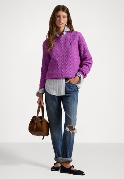 Paarse kabelgebreide trui, wit en blauw gestreept shirt eronder, versleten blauwe spijkerbroek, zwarte instap schoenen, en een bruine handtas.