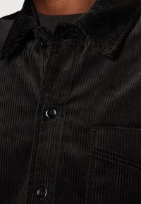 Fekete kordbársony ing gombos gallérral, textúrált bordás anyagból, elülső zsebbel és fekete gombokkal.