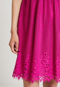 Main à côté d'une robe fuchsia longueur genou avec motif découpé carré et détail en dentelle florale à l'ourlet.