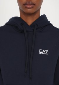 Sudadera azul marino de algodón, con capucha ajustable, bolsillo frontal y un logo blanco de EA7 Emporio Armani en el pecho.