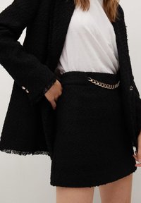 Jupe en tissu texturé noir associée à un blazer assorti, avec un accent chaîne dorée à la taille. T-shirt blanc uni en dessous.